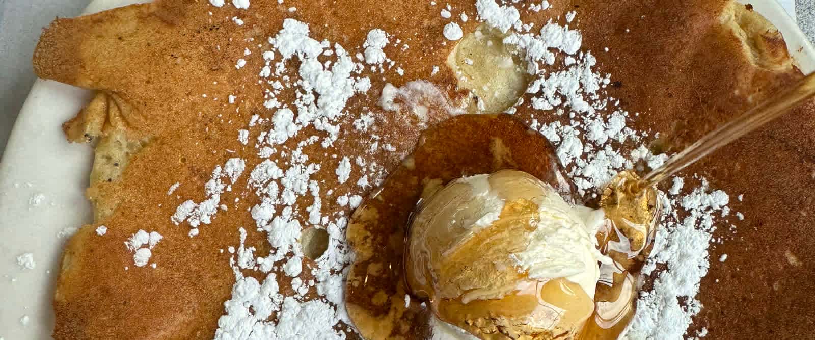 Whipped butter melting into a plate-wide griddle pancake under powdered sugar and pouring syrup